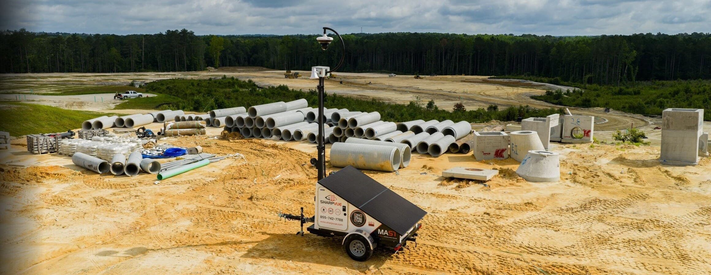 A Sharpvue MAST surveillance trailer deployed on an active construction site, mast raised over stockpiled pipe and graded earth.